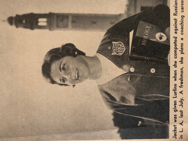 Lurline Hamilton smiles while holding a book and wearing an AAU jacket, standing on LSU’s campus with the Memorial Tower in the background.