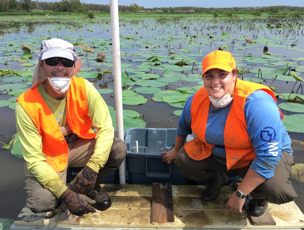 two people in safety vests pose on a dock with lilypads visible in the water behind them