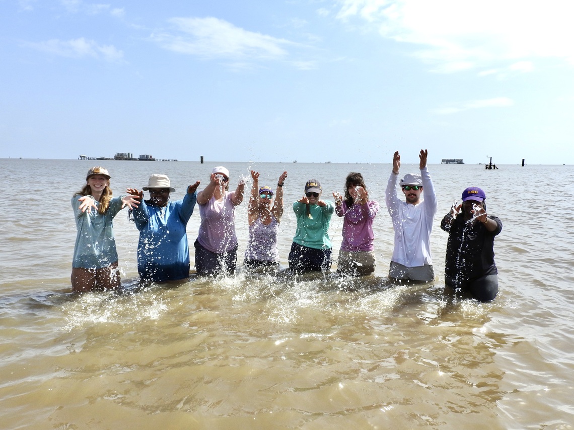 A group of eight people stands in shallow water, splashing and smiling during a coastal field activity.