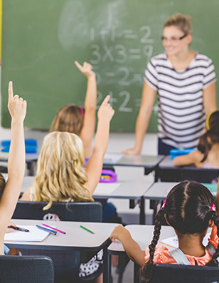 Photo of teacher in front of the classroom with students raising their hands to answer a question.